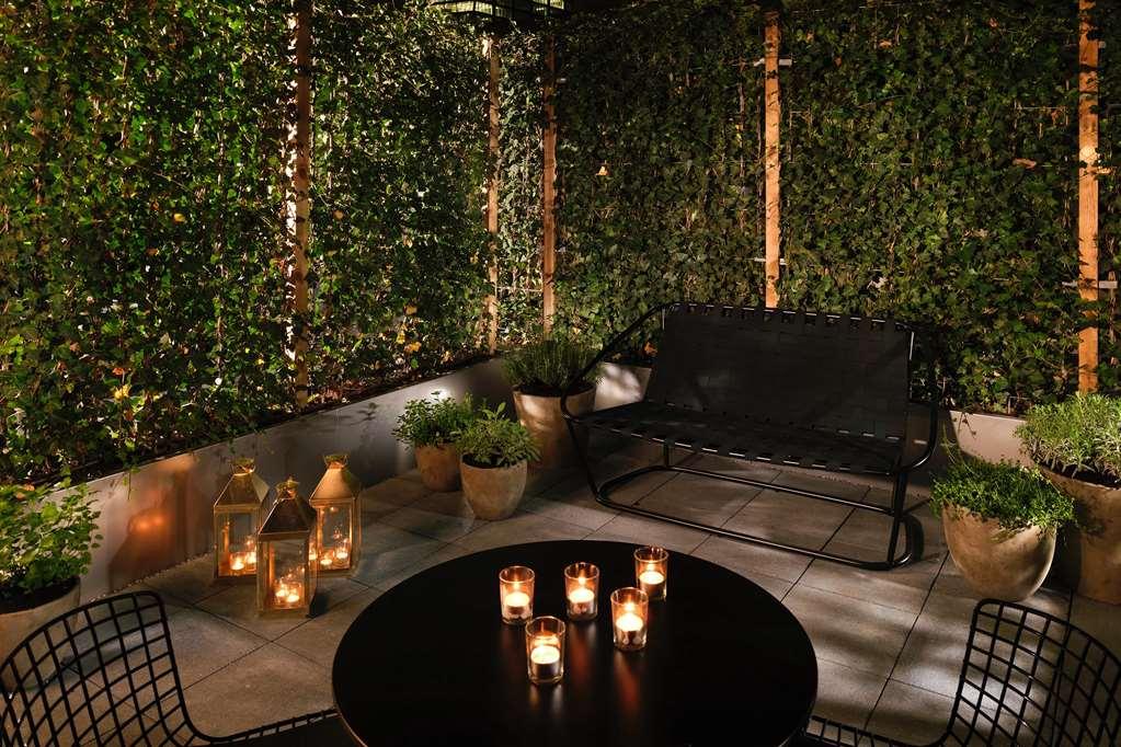 Sea Containers London Hotel Exterior photo The photo depicts a cozy outdoor seating area at night. There is a black metal bench situated against a backdrop of lush greenery, which creates a private and inviting atmosphere. A round black table in the center features several candles, providing 