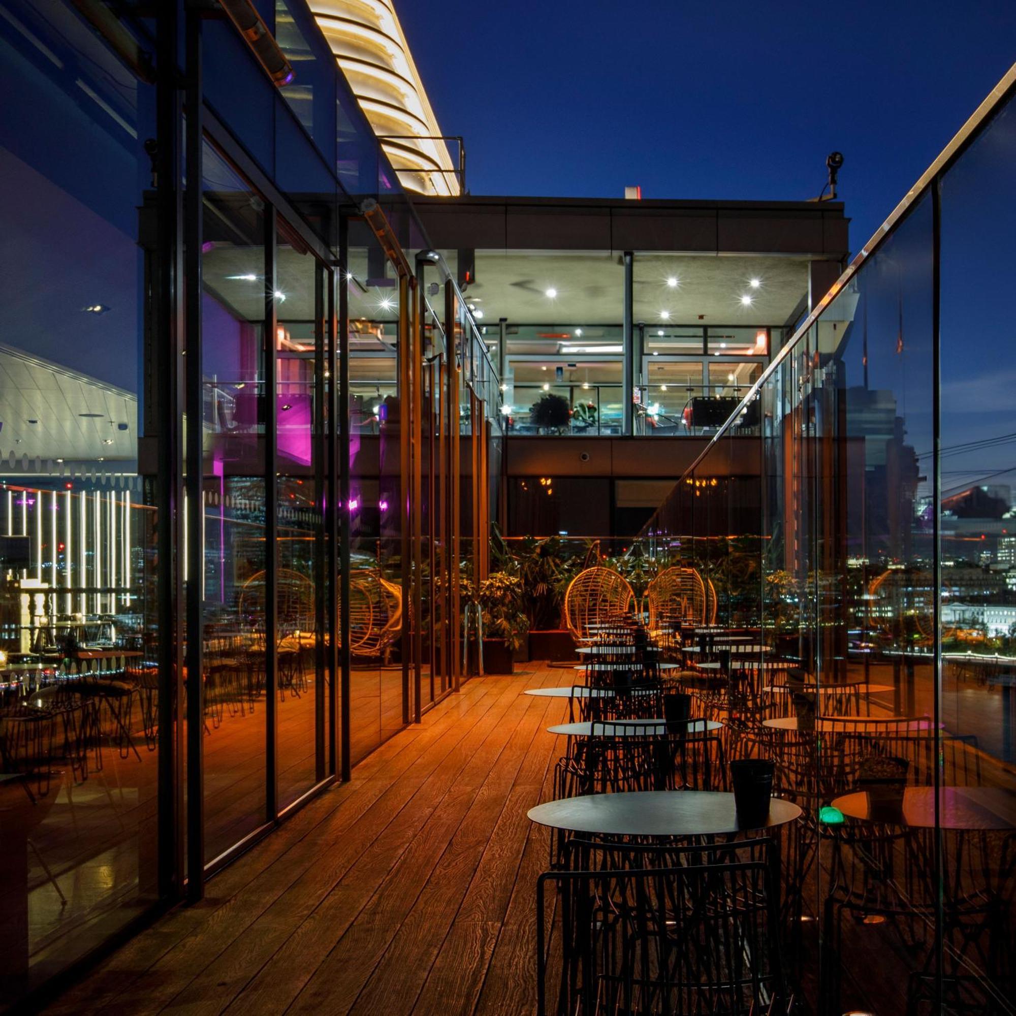 Sea Containers London Hotel Exterior photo The photo depicts a stylish restaurant or lounge area, featuring a modern design with extensive glass walls. It appears to be set on an upper floor, providing a view of the city skyline. The space has a wooden deck and several circular tables surroun