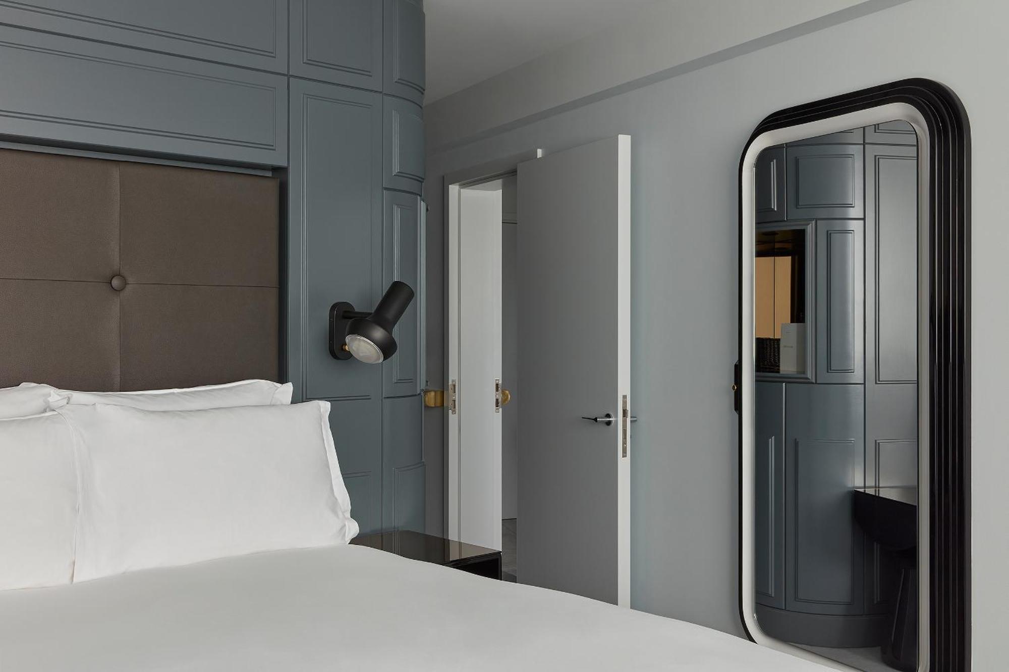 Sea Containers London Hotel Exterior photo The photo shows a modern hotel room or bedroom with a minimalist design. There is a large bed with white linens positioned prominently in the foreground. The walls are painted a muted gray, and some sections have textured paneling. To the right, ther