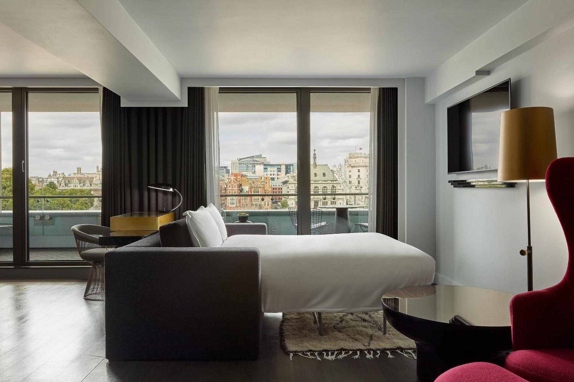 Sea Containers London Hotel Exterior photo The photo shows a modern hotel room with a large window offering a view of a cityscape. The room features a neatly made bed with a white duvet, a stylish gray sofa, and a small round table. There’s a flat-screen television mounted on the wall and a l