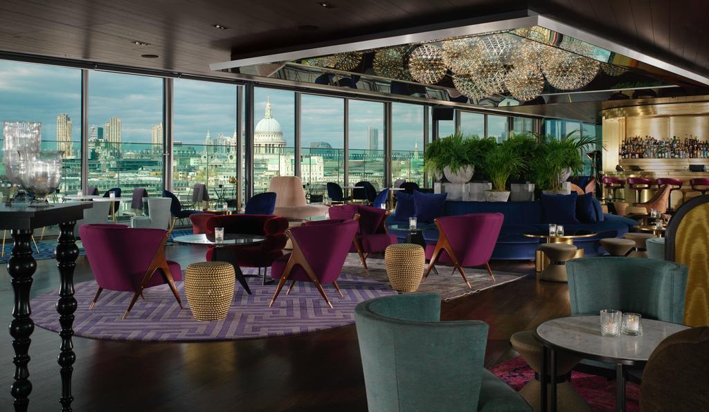 Sea Containers London Hotel Exterior photo The image depicts a stylish interior of a high-rise lounge or bar, featuring large windows that provide a panoramic view of an urban skyline. The decor is modern and elegant, with a mix of seating options, including plush chairs in deep purple and te