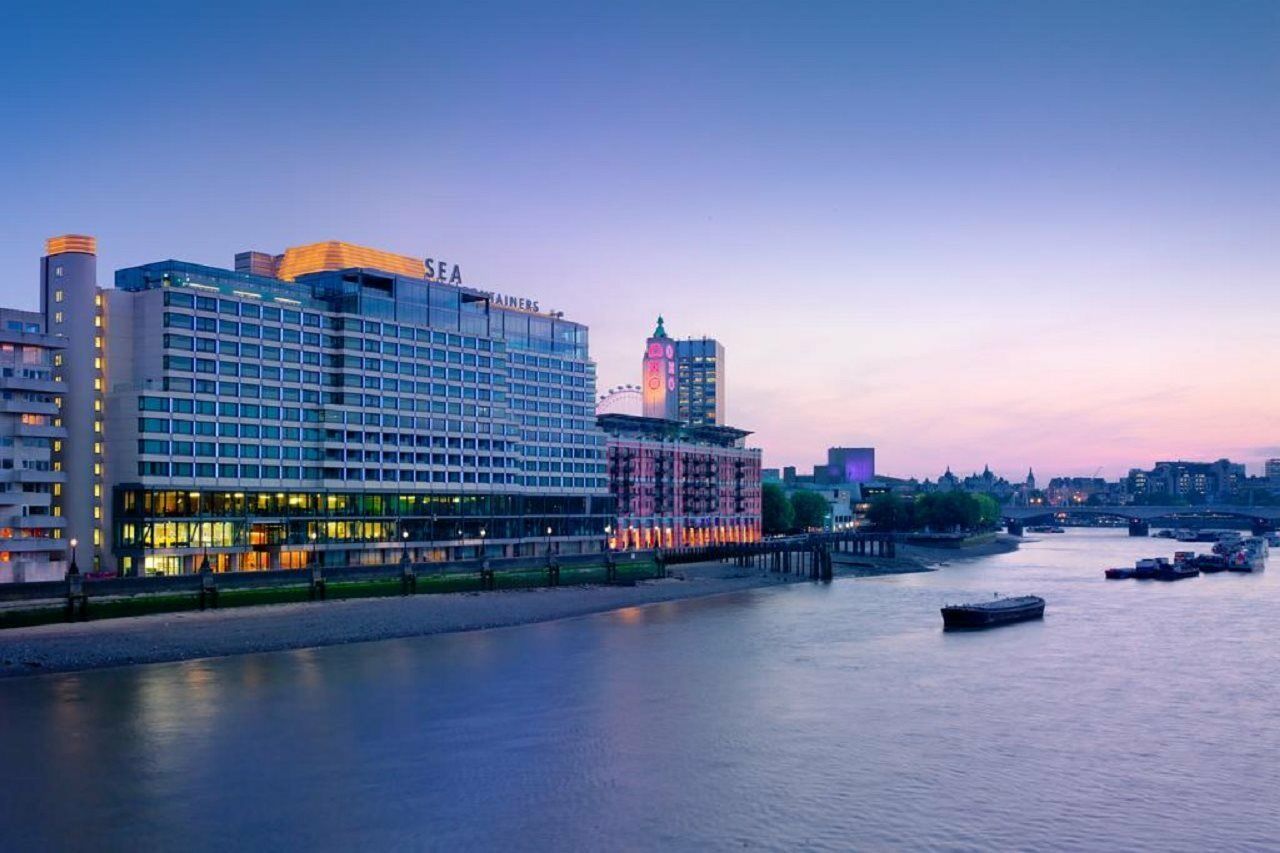 Sea Containers London Hotel Exterior photo The photo shows a waterfront scene featuring a modern hotel or building along a river. The structure has a large façade with lights illuminating it, suggesting it is an upscale destination. The sky is transitioning from daylight to twilight, displayi