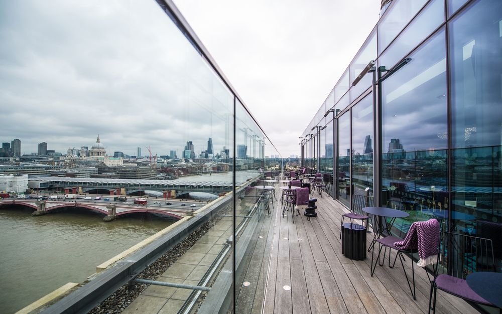 Sea Containers London Hotel Exterior photo The photo depicts a modern balcony or terrace area with a wooden floor and glass railings. It features several outdoor tables and chairs, suggesting it is a space for relaxation or dining. In the background, there are views of a river and city skylin